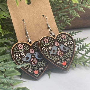 May include: A pair of wooden heart-shaped earrings featuring a floral and bird design. The earrings are painted in shades of blue, green, pink, and red.