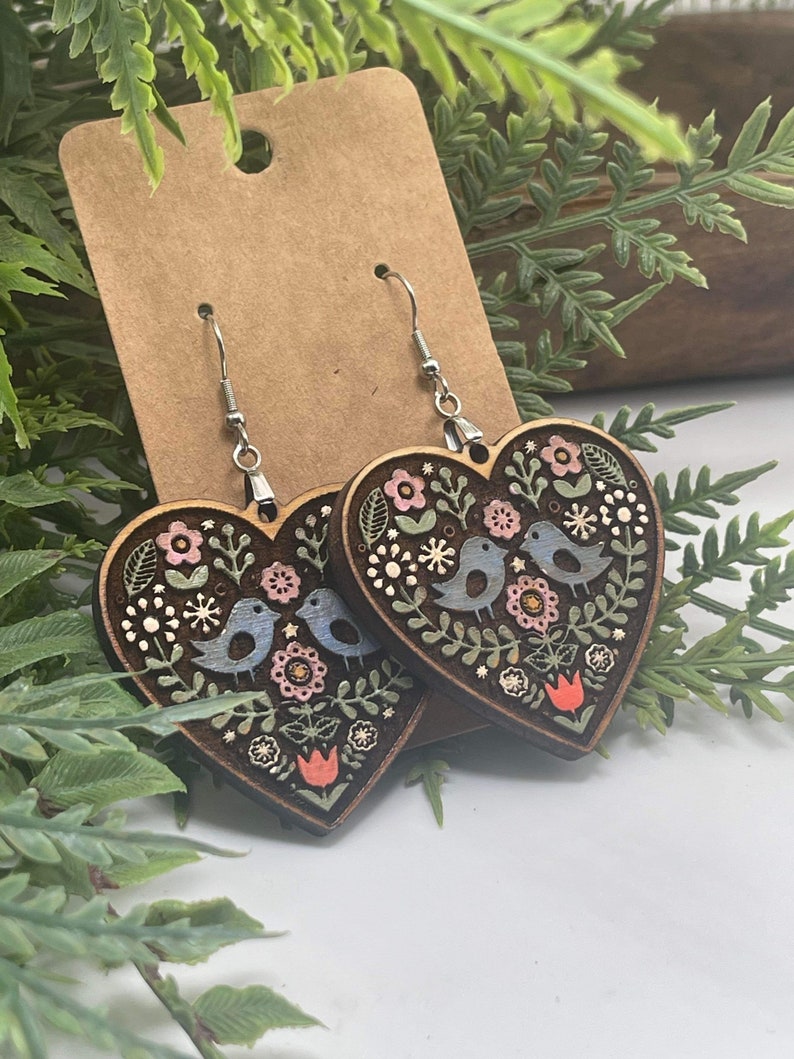 May include: A pair of wooden heart-shaped earrings featuring a floral and bird design. The earrings are painted in shades of blue, green, pink, and red.