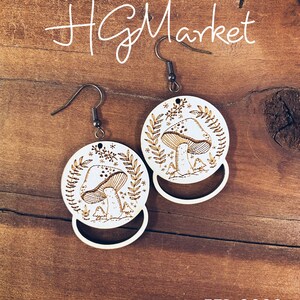 Puede incluir: Un par de pendientes de madera con un diseño de hongo. Los pendientes son blancos con un diseño de hongo marrón y una forma de media luna en la parte inferior. Los pendientes están sobre un fondo de madera.