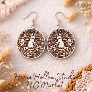 May include: A pair of wooden earrings with a circular design featuring a rabbit surrounded by floral elements. The earrings are brown with white detailing and have silver-colored hooks. The text "Hexie Hollow Studio HG Market" is visible.