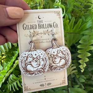 May include: A pair of dangle earrings featuring light-colored wooden pumpkin shapes with dark brown laser-engraved chipmunk illustrations, decorative swirls, and stars. The earrings are attached to a cream-colored card labeled The Gilded Hollow Co.