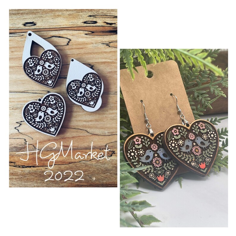 May include: A pair of wooden heart-shaped earrings featuring a floral and bird design. The earrings are painted in shades of brown, pink, blue, and green. The earrings are suspended from silver hooks.