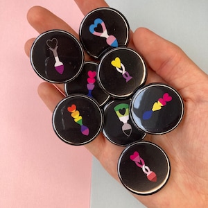 May include: A selection of black, circular badges featuring colourful heart-shaped designs. The badges display pride flag colours and symbols, including rainbow, bisexual, and transgender flags. The badges are held in a hand.