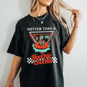K&ouml;nnte beinhalten: Schwarzes T-Shirt mit einem Grafikdruck eines Skeletts in einem Pool-Schwimmring, das ein Getr&auml;nk h&auml;lt. Der Text "Hotter than a Hoochie Coochie" ist auf dem Shirt aufgedruckt.