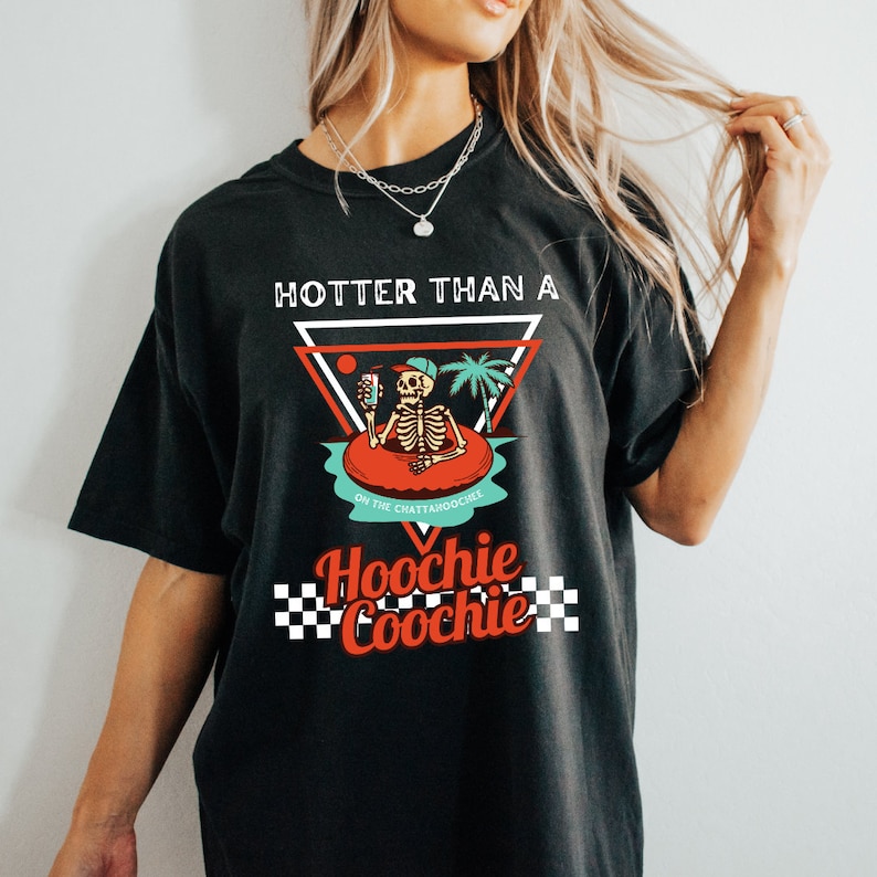K&ouml;nnte beinhalten: Schwarzes T-Shirt mit einem Grafikdruck eines Skeletts in einem Pool-Schwimmring, das ein Getr&auml;nk h&auml;lt. Der Text "Hotter than a Hoochie Coochie" ist auf dem Shirt aufgedruckt.
