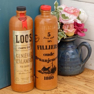 May include: Two vintage glass bottles with cork stoppers and labels. The bottle on the left is labeled "LOOS CARTE OR GENIEVRE DALAMBIO PUR GRAINS". The bottle on the right is labeled "FILLIERS 5 Jaar oud 38% oude graan jenever 1880".