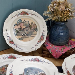 Puede incluir: Un juego de seis platos de cerámica blanca con bordes festoneados y diseños florales. Cada plato presenta una escena diferente de un pueblo europeo, incluyendo una iglesia, casas y un carruaje tirado por caballos.