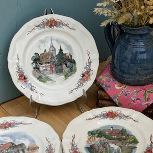 Puede incluir: Un juego de diez platos de cerámica blanca con un borde floral y una escena pintada de un pueblo o campo. Los platos están apilados en dos pilas de cinco.