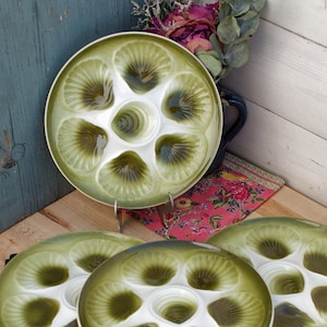 Puede incluir: Juego de cuatro platos de cerámica verde y blanca para ostras, cada uno con seis compartimentos y un pozo central. Los platos tienen un diseño de concha festoneada. Se exhiben sobre una superficie de madera.