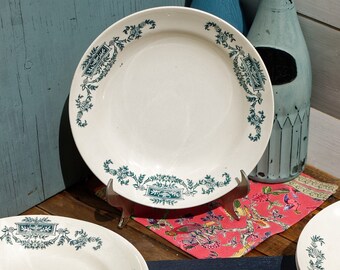 May include: Four white ceramic plates with a green floral design around the rim. The plates are stacked on a blue wooden surface.