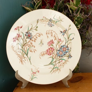 Op de afbeelding: Een crèmekleurige keramische plaat met een bloemenontwerp met roze, blauwe en groene bloemen en tarwearen. De plaat wordt gepresenteerd op een zilveren metalen standaard, die de vintage esthetiek benadrukt. De diameter van de plaat is ongeveer 25 cm.