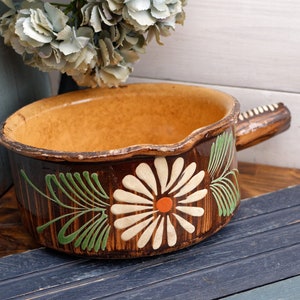 Puede incluir: Una olla de cerámica marrón con un asa de madera. La olla está decorada con diseños florales blancos y verdes y una sola flor naranja.