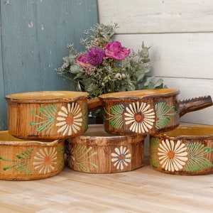 Può includere: Set di cinque teglie da forno in ceramica marrone con motivi di margherite bianche e foglie verdi. Le teglie hanno manici in legno.
