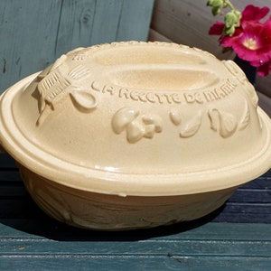 以下が含まれることがあります： 蓋に「La Recette de Mamie」という文字が刻まれた、ベージュの陶器製のパンのドーム型カバー。ドーム型カバーには、葉と花をモチーフにした装飾が施されています。