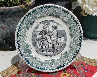 May include: A white ceramic plate with a green and white floral border. The center of the plate features a black and white illustration of two people riding a donkey. The text on the plate reads "Les Misérables en Chemineau".