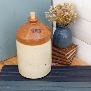 Può includere: Un vaso in ceramica vintage marrone e bianco con un tappo di sughero. Il vaso ha una smaltatura marrone sulla metà superiore e una smaltatura bianca sulla metà inferiore. Il vaso ha le lettere "S.R.D." stampate sul lato.