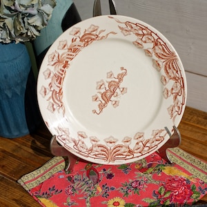 May include: A white ceramic plate with a brown floral design. The plate is on a stand and a red and green floral fabric.