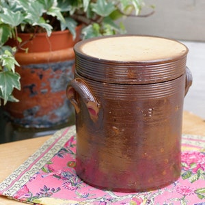 May include: A brown ceramic jar with a lid and two handles. The jar has a glazed finish and is decorated with a series of horizontal lines.