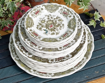 May include: A stack of white ceramic plates with a scalloped edge. The plates feature a floral design in shades of green, pink, and brown. The plates are stacked on a dark blue surface, with greenery in the background.