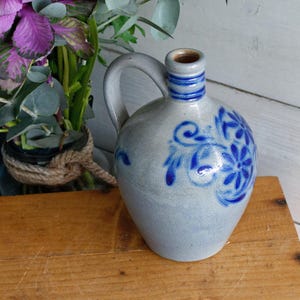 May include: A gray ceramic pitcher with a handle and a blue floral design. The pitcher has a narrow neck with blue stripes and a brown interior. The pitcher is sitting on a wooden surface.