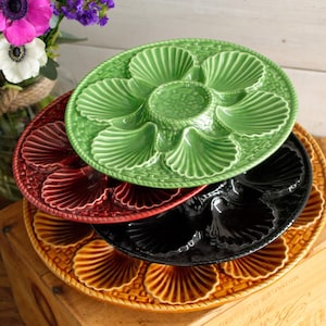 Puede incluir: Una pila de cuatro platos de cerámica para ostras en varios colores: verde, burdeos, negro y ámbar. Cada plato presenta compartimentos en forma de concha y un pozo central. Los platos están dispuestos sobre una superficie de madera.