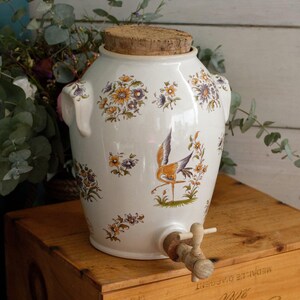 May include: A white ceramic beverage dispenser with a cork lid and a wooden spigot. The exterior features a colorful floral pattern and a central illustration of a crane bird standing on green grass. The vessel has two side handles.