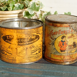 Puede incluir: Dos latas de metal vintage. La lata de la izquierda es amarilla y naranja con texto negro que dice "Encaustique" y "Unigolor Lyon". La lata de la derecha es amarilla y naranja con texto negro que dice "Les Confitures" y "Confituere".