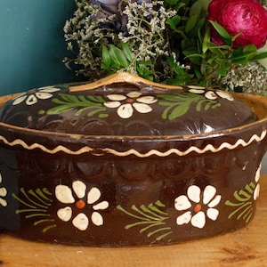 May include: A brown ceramic casserole dish with a lid, decorated with white daisy-like flowers and green leaves. The rim has a wavy, cream-colored design. The dish is on a wooden surface.
