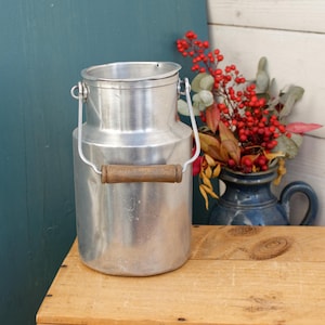 Puede incluir: Una jarra de leche de aluminio plateado con un asa de madera. La jarra está sobre una superficie de madera.