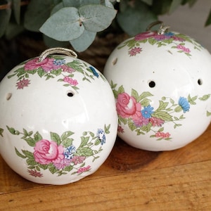 Może przedstawiać: Dwa białe, ceramiczne obiekty w kształcie kuli z kwiatowymi wzorami w kolorze różowym, niebieskim i zielonym. Każda kula ma srebrną pętlę na górze i kilka małych otworów. Kule są umieszczone na drewnianej powierzchni.