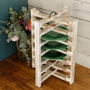 May include: A rustic, white-washed wooden plate rack with a handle, holding several green decorative plates. The rack has a distressed finish and is designed to hold multiple plates. A bouquet of flowers sits in a vase nearby.