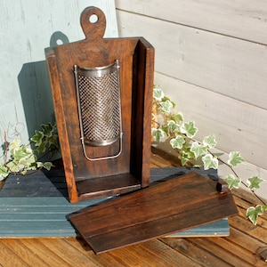 May include: A wooden box with a hinged lid, designed to hold a metal grater. The box is made of dark wood and has a natural finish. The grater is made of stainless steel and has a cylindrical shape.
