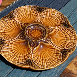 May include: A brown ceramic oyster plate with six individual sections, each shaped like a scallop shell. The plate has a decorative design of raised lines that resemble the texture of a seashell.