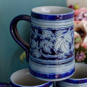 May include: A tall ceramic mug featuring a deep cobalt blue glaze with contrasting light blue horizontal bands and a raised relief pattern of grape leaves and vines wrapping around the center. The mug has a curved handle and a smooth interior.