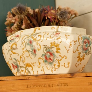 Puede incluir: Jardinera octogonal de cerámica color crema con diseño floral y de volutas en verde azulado, rosa y amarillo. La jardinera está llena de flores secas. La jardinera está sobre una superficie de madera.