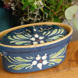 Puede incluir: Una fuente de cerámica azul con tapa, con diseños florales blancos y verdes. El borde y el borde de la tapa son de color beige claro. La fuente está sobre una superficie de madera.