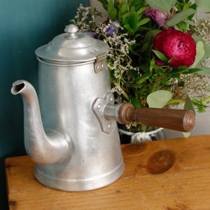Può includere: Una caffettiera vintage in alluminio con manico e coperchio in legno. La caffettiera ha un beccuccio e un supporto per manico decorativo. Lo sfondo include un bouquet di fiori e una superficie in legno.