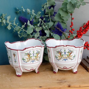 Puede incluir: Dos macetas de cerámica blanca con ribetes rojos y dorados y diseños florales. Las macetas tienen tres patas cada una y están sobre una superficie de madera.