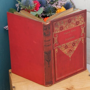 May include: A red hardcover book with gold lettering on the spine and front cover. The title of the book is "Walter Scott Ivanhoe".