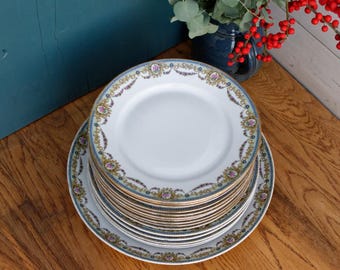 Op de afbeelding: Een stapel witte porseleinen dinerborden met een gouden en blauw bloemenpatroon. De borden zijn gestapeld op een houten oppervlak.