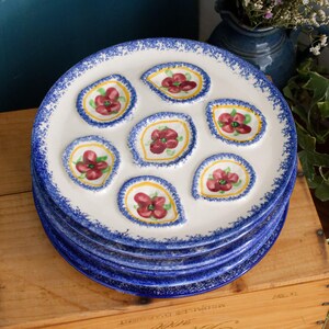 May include: A stack of white ceramic plates with blue rims and floral designs. Each plate features seven indentations, each holding a red flower with yellow accents. The plates are stacked on a wooden surface.