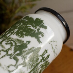 Peut inclure: Pot en céramique blanc avec un bord noir, orné d'un motif de toile de Jouy vert représentant une personne à cheval, des arbres et des lampadaires. Le pot a une esthétique classique et vintage.
