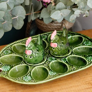 Op de afbeelding: Een groene keramische eierhouder met een bloemmotief en twee zout- en pepervaatjes in de vorm van kippen. De eierhouder heeft ovale compartimenten voor eieren. De kippen hebben roze accenten op hun kammen en lellen.