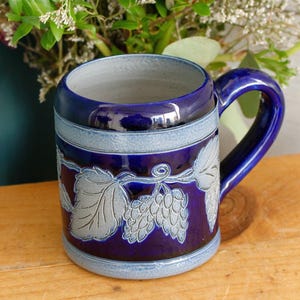 May include: A dark blue ceramic beer stein with a handle. The stein features a light blue band around the top and bottom, and a decorative relief of grape vines and leaves. The interior is a light gray color.