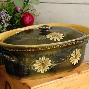 Peut inclure: Plat à four ovale en céramique vert foncé avec couvercle. Décoré de marguerites peintes à la main et d'un bord jaune. Le couvercle a un petit bouton rond. Le plat est posé sur une caisse en bois.