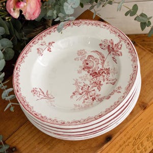 Puede incluir: Una pila de platos de cerámica blancos con un diseño floral rojo. Los platos tienen un borde rojo decorativo y un patrón floral detallado en el centro. Los platos están apilados sobre una superficie de madera.