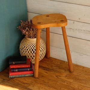 Puede incluir: Un pequeño taburete de madera de tres patas con un asiento redondeado. El taburete es de madera de color claro y tiene un acabado natural. Las patas están ligeramente anguladas. Un jarrón de mimbre con flores secas y una pila de libros están en el fondo.