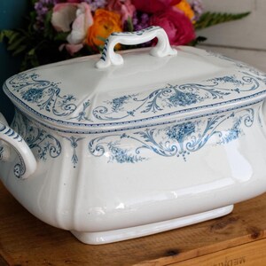 Puede incluir: Una sopera rectangular de cerámica blanca con patrones florales y de volutas en azul. La pieza incluye una tapa a juego con un asa central y dos asas laterales, todas decoradas con detalles en azul sobre una superficie blanca brillante.