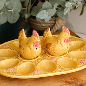 Puede incluir: Un huevera de cerámica amarilla con dos saleros y pimenteros con forma de gallina. El huevera tiene espacios para huevos y está decorada con detalles florales rosas. Las gallinas tienen crestas y picos rosas.