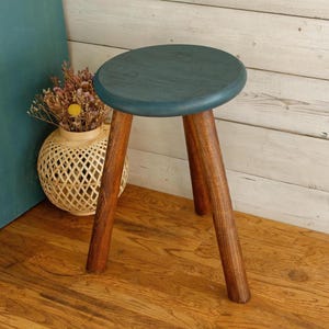 Puede incluir: Un pequeño taburete de madera con un asiento redondo de color verde azulado y tres patas marrones. El taburete está sobre un suelo de madera. Un jarrón decorativo con flores secas está al lado.